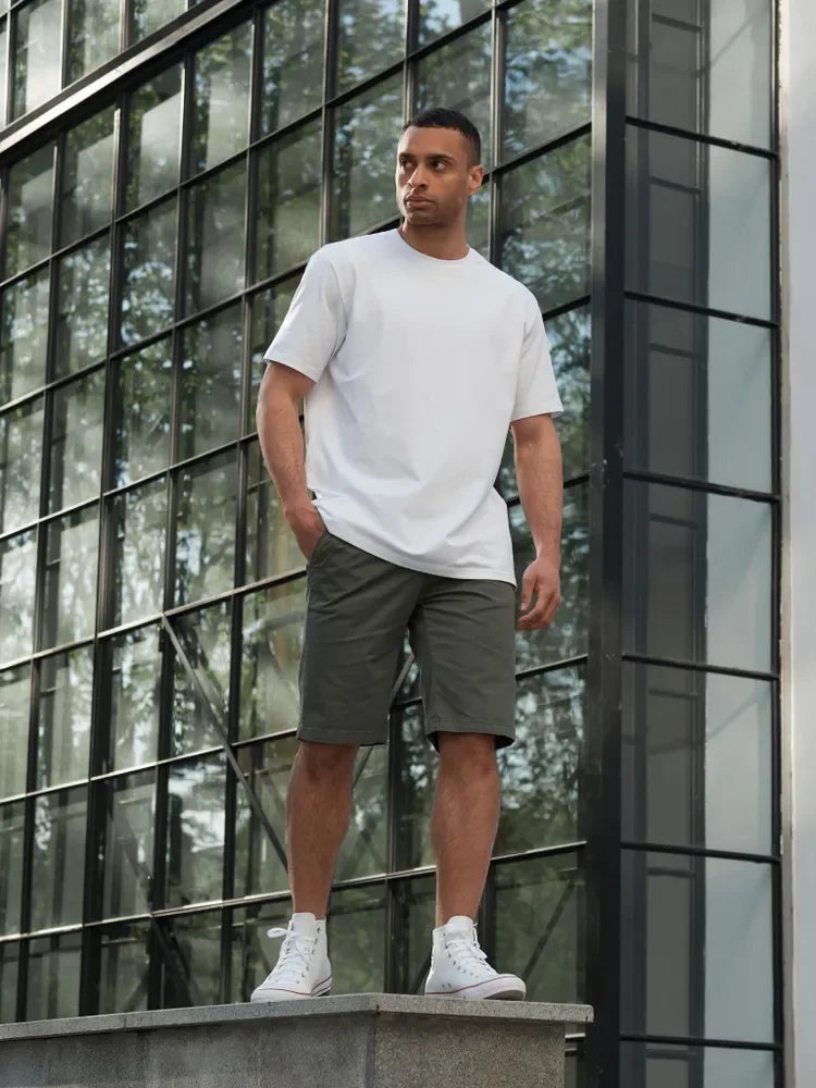 Man wearing a white t-shirt and gray shorts standing in front of a modern glass building.