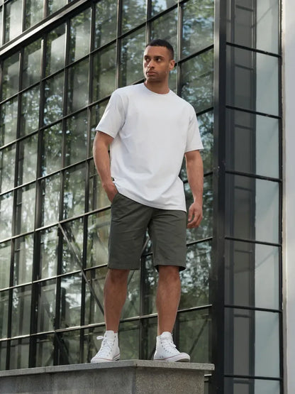 Man wearing a white t-shirt and gray shorts standing in front of a modern glass building.