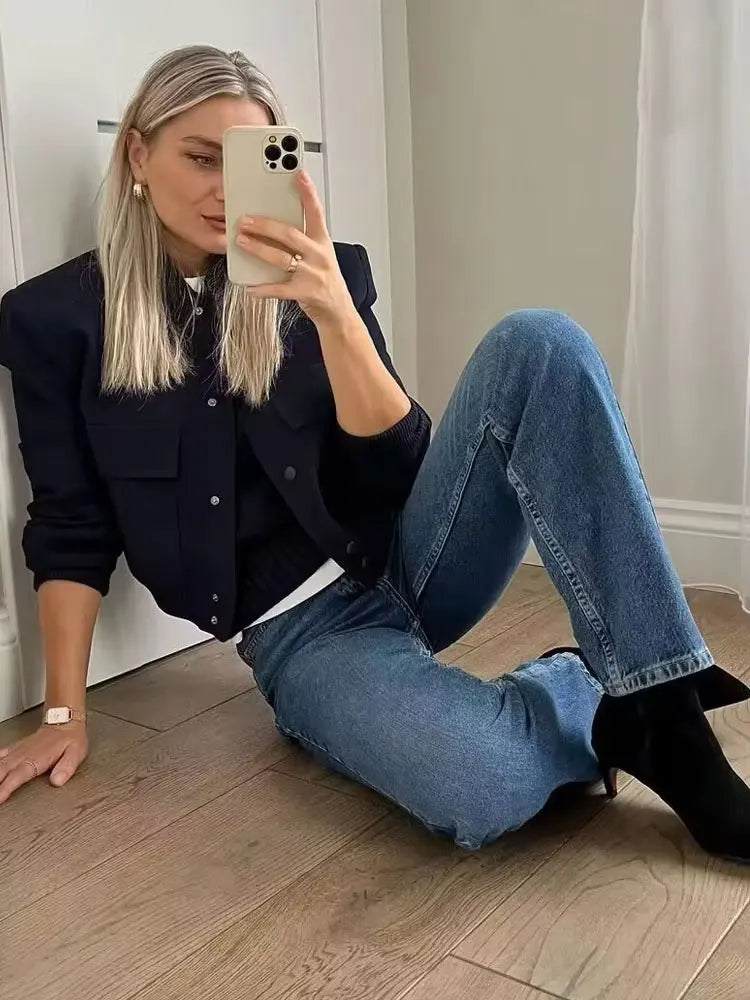 Person sitting on the floor taking a mirror selfie wearing a black jacket and blue jeans.
