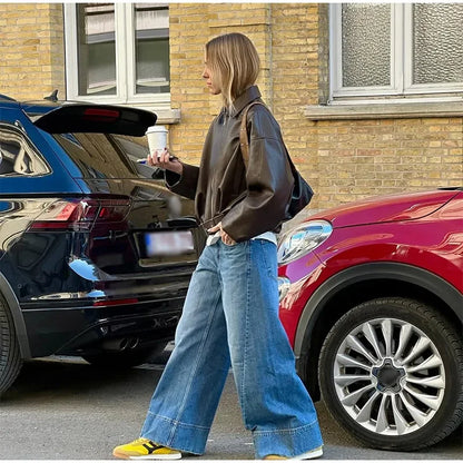 Person in brown leather jacket and blue jeans standing next to a black car with a red car in the background.