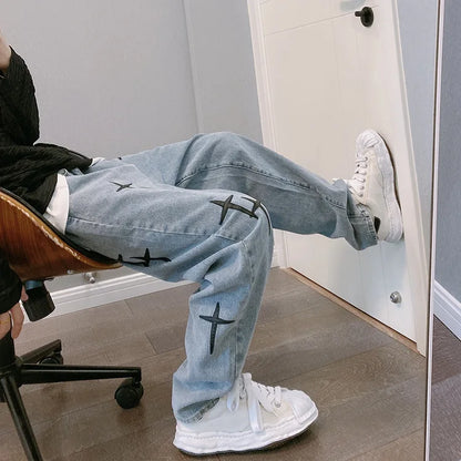 Person wearing light blue jeans with black cross designs and white sneakers sitting on a chair.