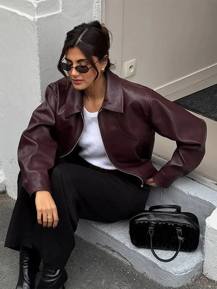 Woman wearing a brown leather jacket sitting on steps with a black handbag beside her.