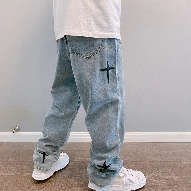 Light blue jeans with black cross designs worn by a person against a plain wall.