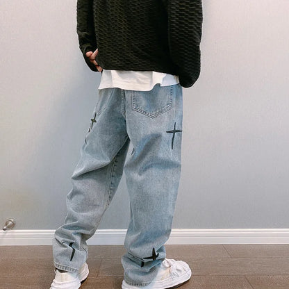 Person wearing light blue jeans with black cross designs, a black sweater, and white sneakers against a plain wall.