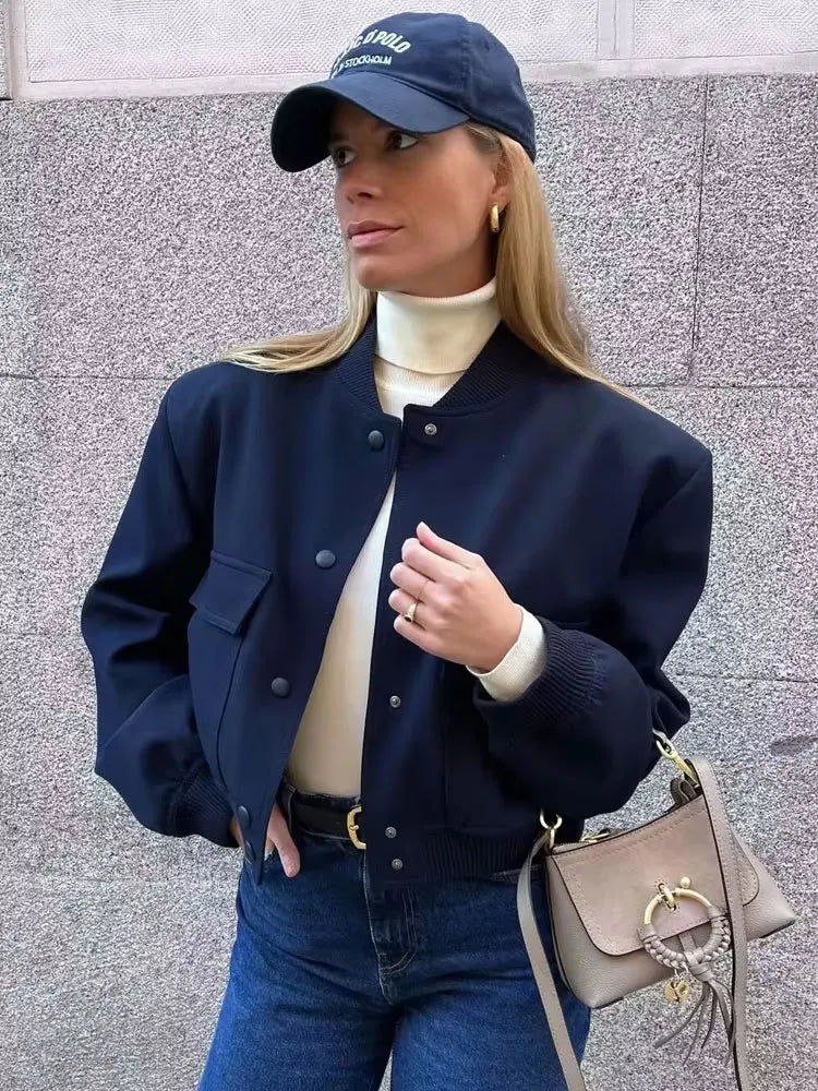 Woman wearing a navy jacket, white turtleneck, blue jeans, and navy cap against a gray wall.