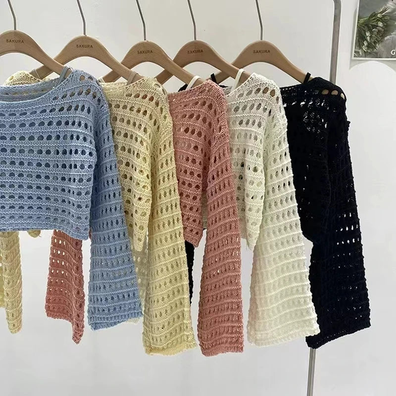 Five crocheted sweaters in blue, yellow, pink, white, and black hanging on wooden hangers against a white background.