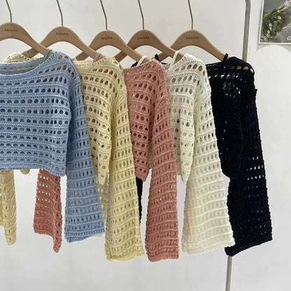 Five crocheted sweaters in blue, yellow, pink, white, and black hanging on wooden hangers against a white background.