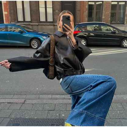 Person wearing a black leather jacket and blue jeans taking a mirror selfie on a city street.
